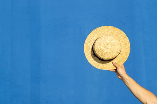 A man's hand holding a straw hat on a blue background. Summer, vacation concept.