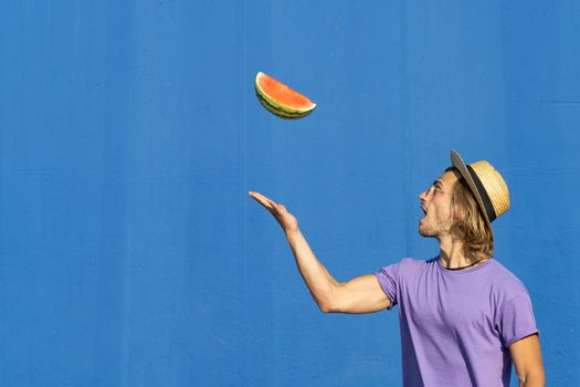 Young man in hat throwing a piece of watermelon on a blue background. Summer, diet and weight loss concept.