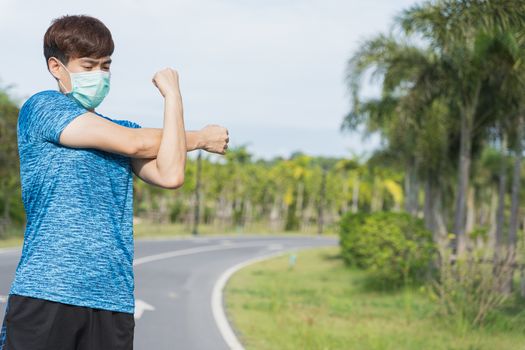 Young male wearing medical mask and stretching his arms before workout training session at the park. Stay in fit during quarantine in the days of the Corona Virus or Covid-19