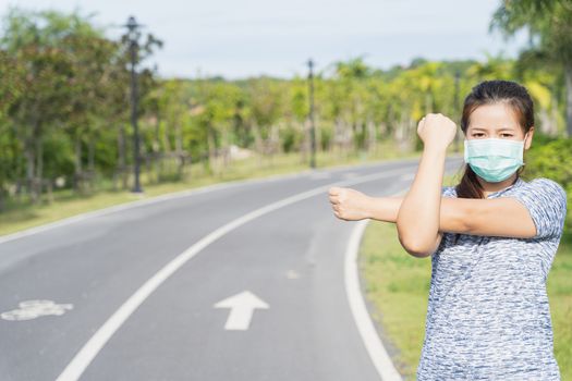 Young female wearing medical mask and stretching his arms before workout training session at the park. Stay in fit during quarantine in the days of the Corona Virus or Covid-19