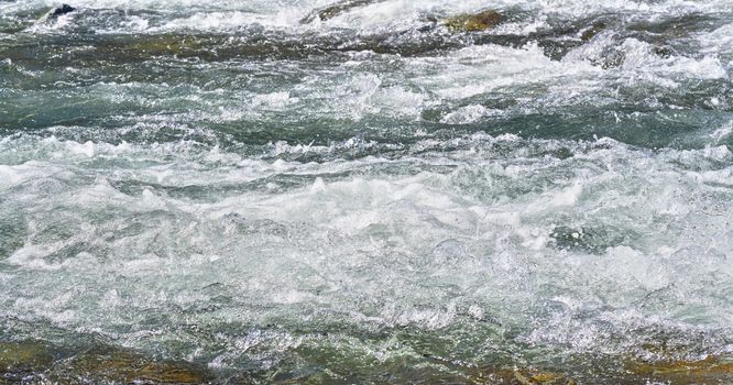 Rapid spring river flowing over rocks on sunny day, forming white water waves, closeup detail - abstract nature background.