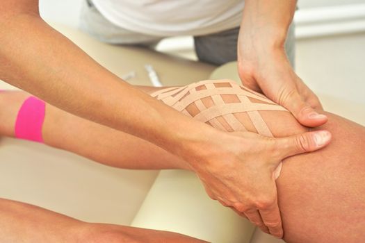 Young female physiotherapist applying kinesiology tape and massaging knee of her patient, closeup detail on hands.