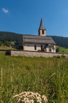 South Tyrolean mountain church under a blue sky with a single white cloud, mountain landscape with meadows and pines