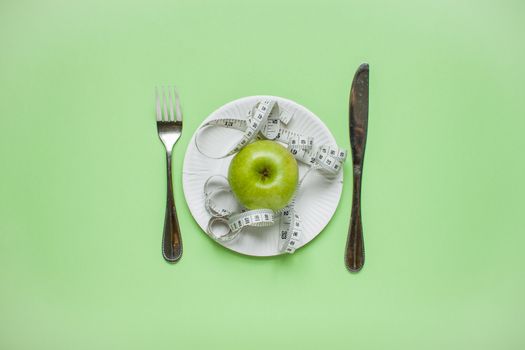 Diet and healthy eating concept. Top view of weightloss. Green apple and measuring tape on a plate, knife with a fork. Green background