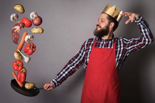 Cooking man concept, king of kitchen,bearded man in checked shirt, drop up meat and vegetables from a pan, studio shot on gray background