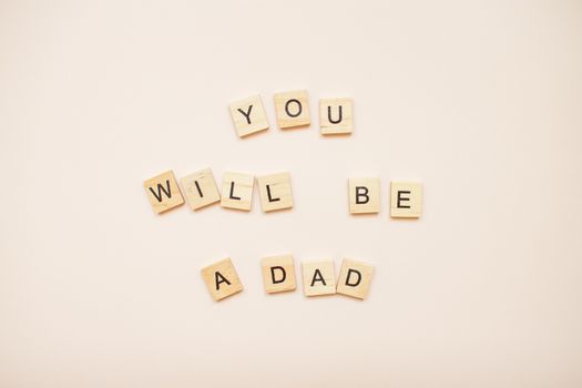 The inscription "you will be a dad" made of wooden blocks on a light pink background.
