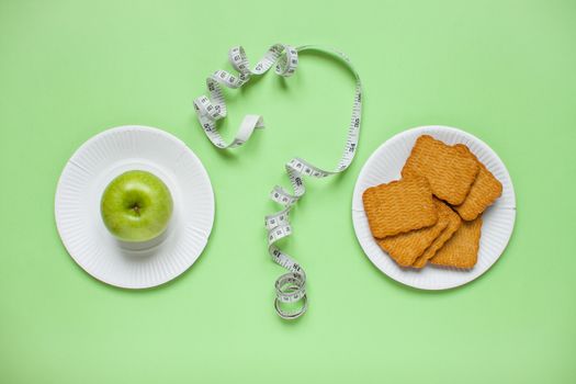 Diet and healthy eating concept. Top view of weightloss. The choice between a green apple and cookies. Question mark from a measuring tape. Green background