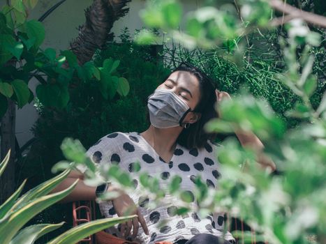 Beautiful Asian woman wearing protective face mask and polka dot shirt closing her eyes sitting on the red chair in the green garden, new normal lifestyle concept.