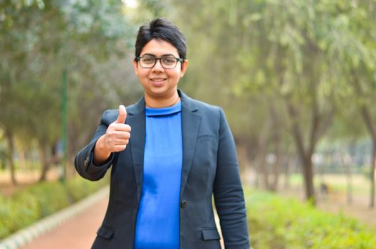 Portrait of a confident young Indian Corporate professional woman with short hair showing thumbs up sign in an outdoor setting wearing a black business / formal suit