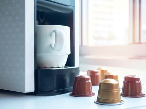 White ceramic coffee cup on coffee machine and espresso coffee capsules on white table near the window with sunlight in the morning.