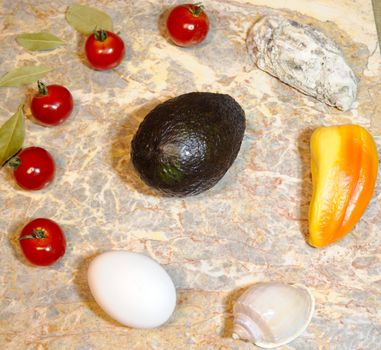 vegetables, fruits, seashells and eggs on a marble surface: cherry tomatoes, bell peppers, bay leaves, oyster, seashell, avocado and chicken egg