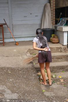 Woman cleaning service with broom stick and dustpan clean up the road. Balinese girl sweeps garbage and dry leaves in front of the house. Bali, Indonesia.