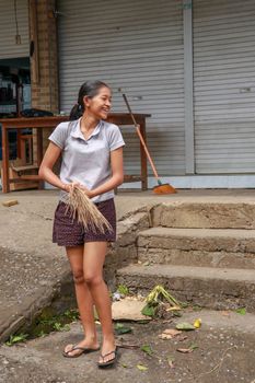 Woman cleaning service with broom stick and dustpan clean up the road. Balinese girl sweeps garbage and dry leaves in front of the house. Bali, Indonesia.