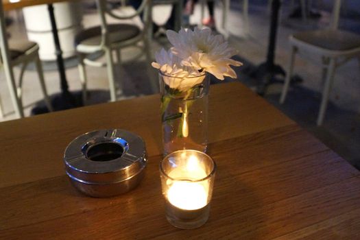 lighted candle, chrysanthemum flower and ashtray on a wooden table of a street cafe in the evening.