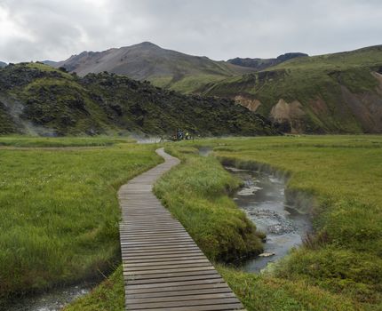 Wooden path to natural hot spring with group of tourist people relaxing in a thermal baths in Landmannalaugar camp site, Iceland. Grass meadow, lava fields and mountains in background.