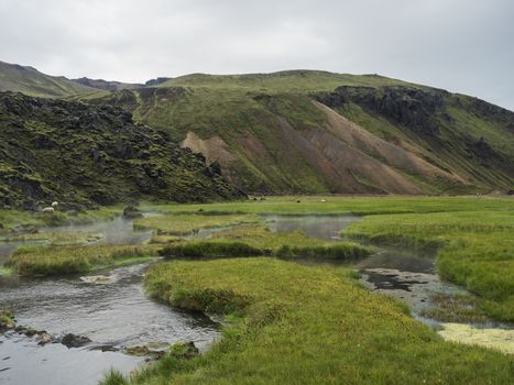 View on geothermal area with natural hot spring, thermal baths in Landmannalaugar camp site, Iceland. Grass meadow, lava fields and mountains in background.