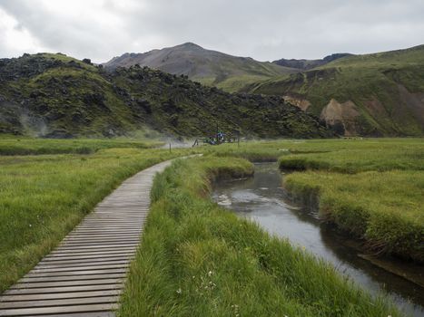 Wooden path to natural hot spring with group of tourist people relaxing in a thermal baths in Landmannalaugar camp site, Iceland. Grass meadow, lava fields and mountains in background.