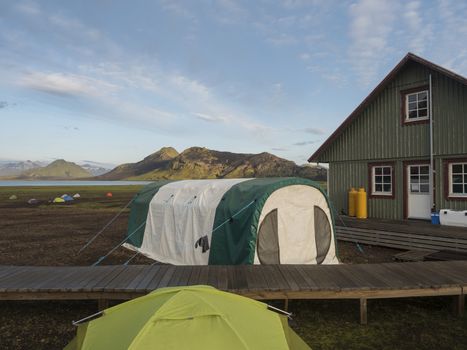 Colorful tents and green tourist hut at Alftavatn camping site with blue lake and green hills and glacier in the background. Landscape of the Fjallabak Nature Reserve in the Highlands of Iceland part of famous Laugavegur hiking trail