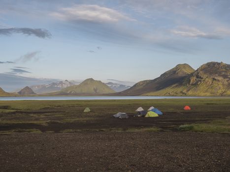 Colorful tents at camping site on blue Alftavatn lake with green hills and glacier in the otherwordly beautiful landscape of the Fjallabak Nature Reserve in the Highlands of Iceland. Part of famous Laugavegur hiking trail.