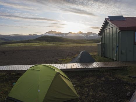 Colorful tents and green tourist hut at Alftavatn camping site with snow covered mountains and green hills and glacier in the background. Landscape of the Fjallabak Nature Reserve in the Highlands of Iceland part of famous Laugavegur hiking trail. Sunrise