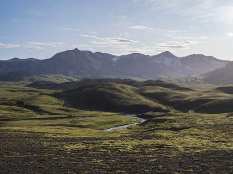 Volcanic landscape with blue river, snow covered mountains, green hills and lava gravel ground covered by lush moss. Fjallabak Nature Reserve in the Highlands of Iceland.