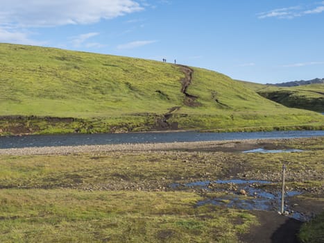 Group of hikers preparing to ford across the blue river stream at Laugavegur hiking trail. Landscape with green hills, meadow and lush moss. Fjallabak Nature Reserve, Iceland. Summer blue sky.