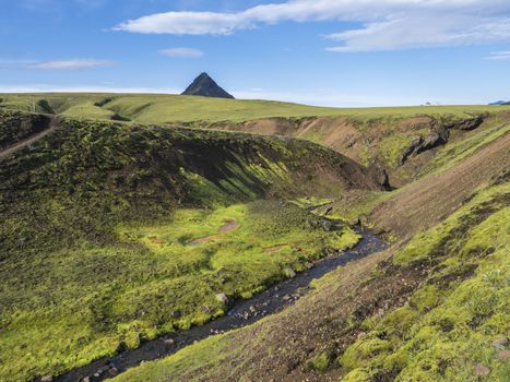 Panoramic volcanic landscape of green Storasula mountain with lush moss and blue creek water between Emstrur and Alftavatn camping sites on Laugavegur trek in area of Fjallabak Nature Reserve, Iceland.