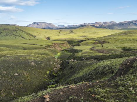 Panoramic volcanic landscape of green Storasula mountain with lush moss and blue creek water between Emstrur and Alftavatn camping sites on Laugavegur trek in area of Fjallabak Nature Reserve, Iceland.