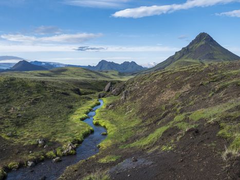 Panoramic volcanic landscape of green Storasula mountain with lush moss and blue creek water between Emstrur and Alftavatn camping sites on Laugavegur trek in area of Fjallabak Nature Reserve, Iceland.