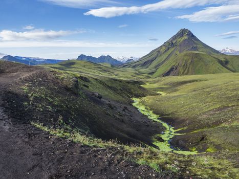 Panoramic volcanic landscape of green Storasula mountain with lush moss and blue creek water between Emstrur and Alftavatn camping sites on Laugavegur trek in area of Fjallabak Nature Reserve, Iceland.