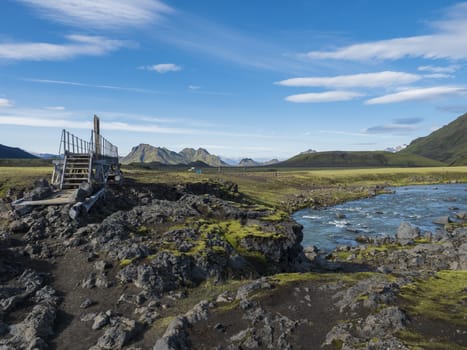 Landscape with wooden footbridge over a blue glacier river at laugavegur hiking trail in iceland. Lava formation and green hills and mountains, summer blue sky background.