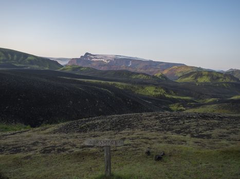 wooden tourist post with sign Thosmork at Botnar campsite at Iceland on Laugavegur hiking trail, green valley in volcanic landscape among lava fields with view on Myrdalsjokull glacier. Early morning sunrise view
