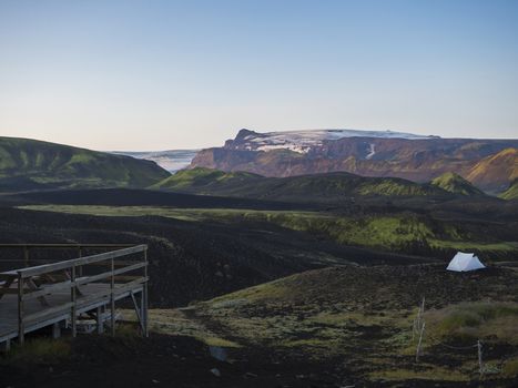 Wooden terrace and white tent in Botnar campsite at Iceland on Laugavegur hiking trail, green valley in volcanic landscape with view on Myrdalsjokull glacier. Early morning sunrise pink light.