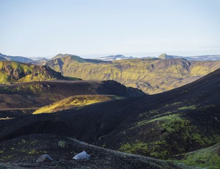Three tents alone in Botnar campsite at Iceland on Laugavegur hiking trail, green valley in volcanic landscape among lava fields with view on Myrdalsjokull glacier. Early morning sunrise view.