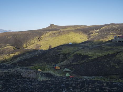 Colorful tents and Botnar mountain hut at Iceland on Laugavegur hiking trail, green valley in volcanic landscape among lava fields with view on Myrdalsjokull glacier. Early morning sunrise view