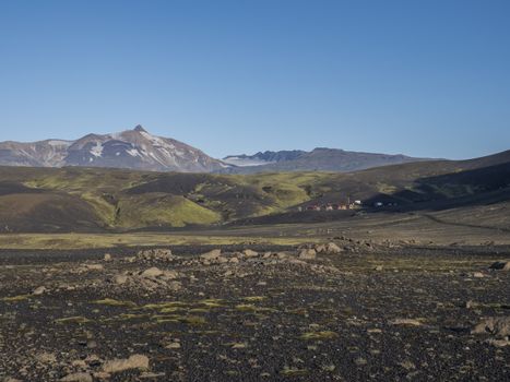 View on Botnar mountain hut and campsite at Iceland on Laugavegur hiking trail, green valley in volcanic landscape among lava fields with view on Myrdalsjokull glacier. Early morning, summer blue sky
