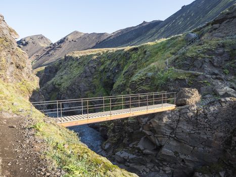 Landscape with wooden footbridge over a blue glacier river at laugavegur hiking trail in iceland. Lava formation and green hills and mountains, summer blue sky background.