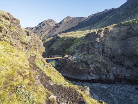 Landscape with wooden footbridge over a blue glacier river at laugavegur hiking trail in iceland. Lava formation and green hills and mountains, summer blue sky background.
