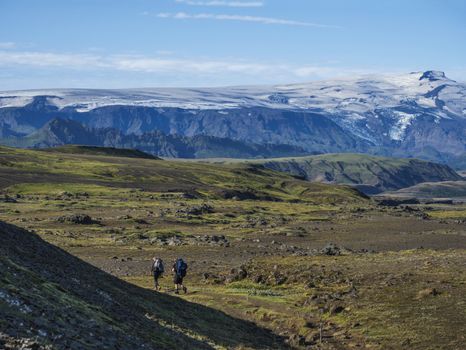 Couple of two young hikers at Laugavegur hiking trail with eyjafjallajokull volcano and glacier tongue, blue river stream and green hills. Fjallabak Nature Reserve, Iceland. Summer blue sky.