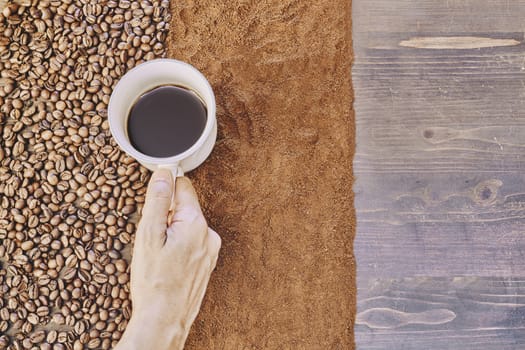 Top view of a person holding a cup of coffee with roasted and ground coffee beans on the table