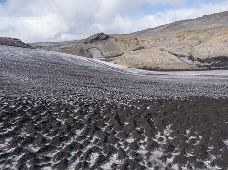 Landscape of Fimmvorduhals hiking trail with close up snow filed covered by black lava ash dunes and volcanic mountains. Summer blue sky.
