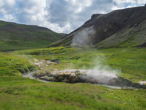 Reykjadalur valley with hot springs river and pool with lush green grass meadow and hills with geothermal steam. South Iceland near Hveragerdi city. Summer sunny morning, blue sky