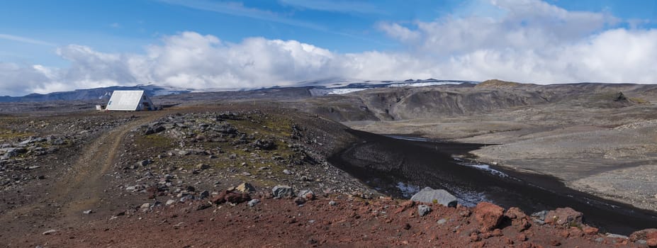 Panoramic red and black volcanic Iceland landscape with mountain hut shelter Baldvinsskali at Fimmvorduhals pass on hiking trail and Eyjafjallajokull glacier volacano peak. South Iceland, Summer blue sky.