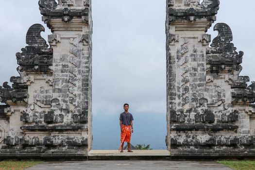 A young man in traditional clothes passes through the gate looking back. Ancient gate in Pure Lempuyan, Bali, Indonesia.