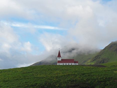 lonely red white church in vik in iceland green grass hills and low clouds