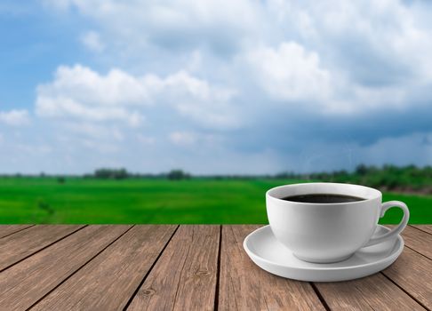 White cup of hot coffee on a wooden table with fields and the sky as a blurred background.
