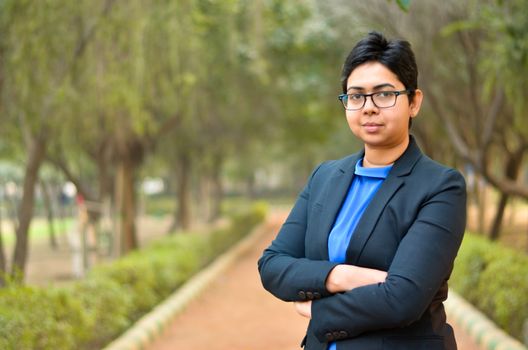 Closeup portrait of a confident young Indian Corporate professional woman with short hair and spectacles, crossed folded hands in an outdoor setting wearing a black business / formal suit