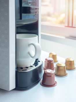 White ceramic coffee cup on coffee machine and espresso coffee capsules on white table near the window with sunlight in the morning, vertical style.