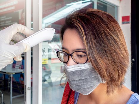 Body Temperature check. Hand holding electronic infrared thermometer on woman forehead in front of supermarket, Coronavirus (COVID-19) prevention.