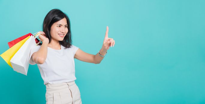 Portrait happy Asian beautiful young woman standing wear white t-shirt, She holding shopping bags multi color on hand and touch air by finger, shoot photo in a studio on a blue background